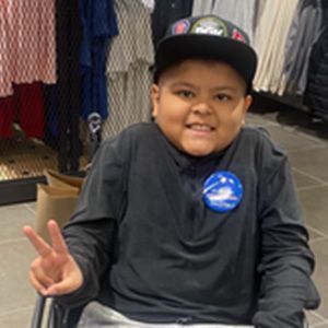 Boy in a wheelchair wearing a cap and black shirt gives a peace sign, smiling in a store.