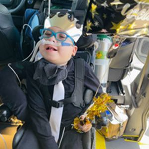 Boy in formal attire, smiling with glasses and medical equipment, holding a gold decoration, with a celebratory balloon.