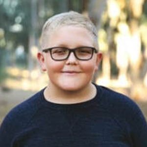 Young person with glasses smiling, wearing a navy blue sweater outdoors.