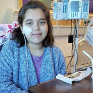 Young girl in hospital bed, wearing a blue sweater and a nasal cannula, smiling.