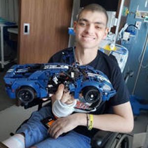 Man in wheelchair holds a blue Lego car, smiling. He wears a black shirt, in a hospital room.