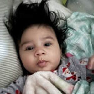 Baby with messy dark hair, puckered lips, looking at the camera. Wearing patterned pajamas in bed.