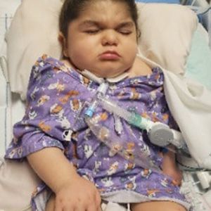 A child with a breathing tube in a hospital bed, wearing purple pajamas, with a sad expression.