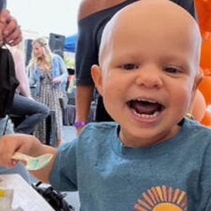 Smiling toddler with shaved head holding food, blue shirt, outdoors.