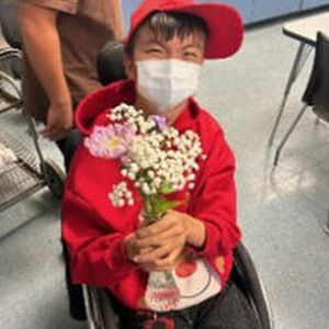 Person in wheelchair, wearing red hoodie/hat/mask, holding flowers, smiling.