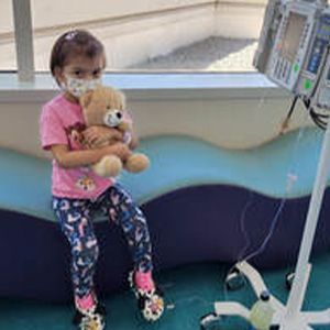 Young girl wearing a mask holds a teddy bear, sitting near medical equipment.