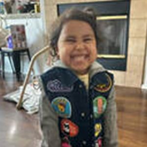 Smiling child wearing a vest with colorful patches stands indoors in front of a fireplace.