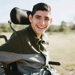 Smiling young man in a wheelchair outdoors, wearing a green and gray shirt.