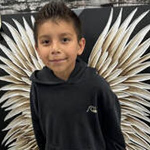 Boy with dark hair in black hoodie stands in front of large angel wings, smiling.