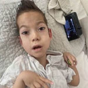 Boy with brown hair and light skin, lying in bed, wearing a light colored shirt, with an electronic device.