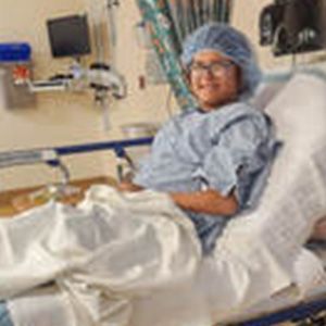 Person wearing a surgical cap and gown smiles while lying in a hospital bed.