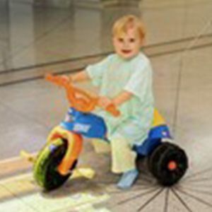 A young child with blonde hair rides a blue, yellow and orange tricycle, smiling.