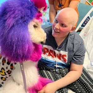 A smiling girl with a shaved head looks at a poodle with colorful fur in a hospital room.