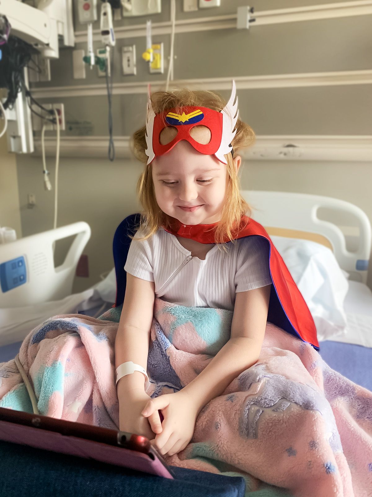 Girl in a superhero costume sits on a hospital bed, smiling while looking at a tablet.