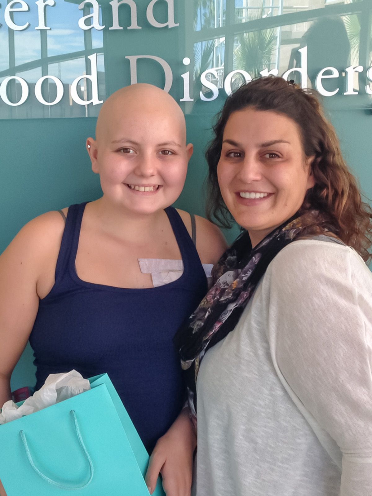 A young person with a bald head smiles with a woman, both are near a hospital sign that says 