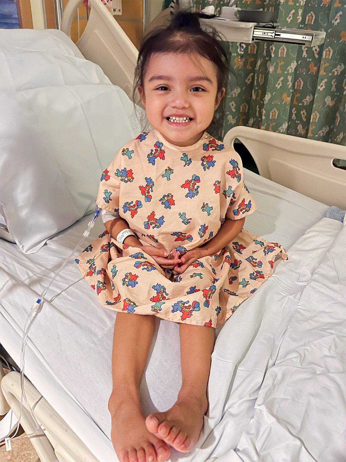 Smiling young girl in a hospital bed wearing a patterned gown.