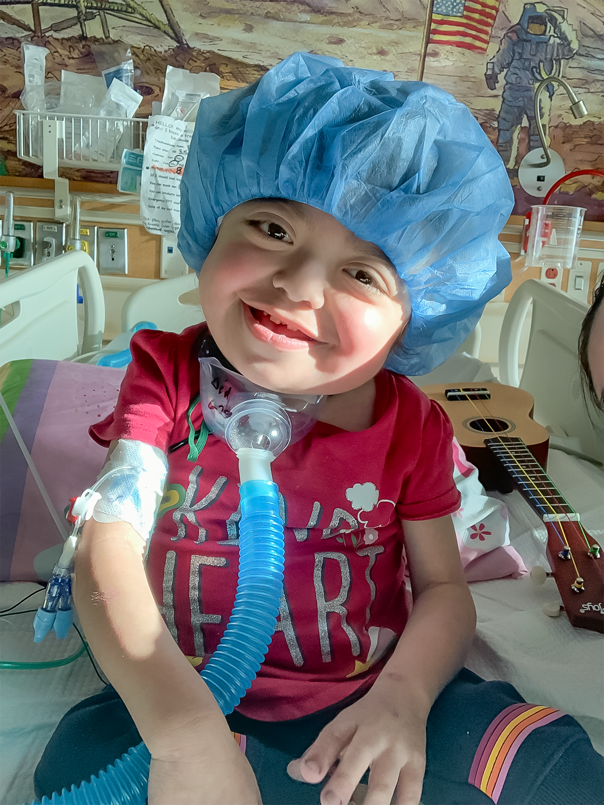 Smiling child wearing a surgical cap and medical tubing, sitting in a hospital bed.