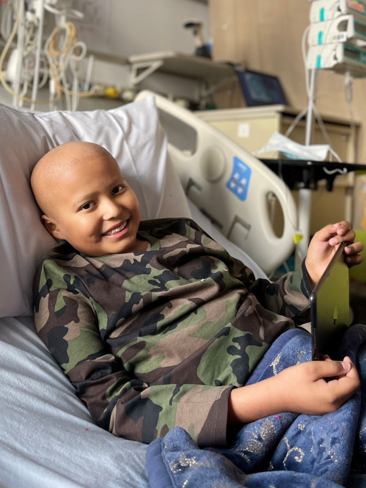 Smiling child with bald head in hospital bed wearing camouflage shirt.