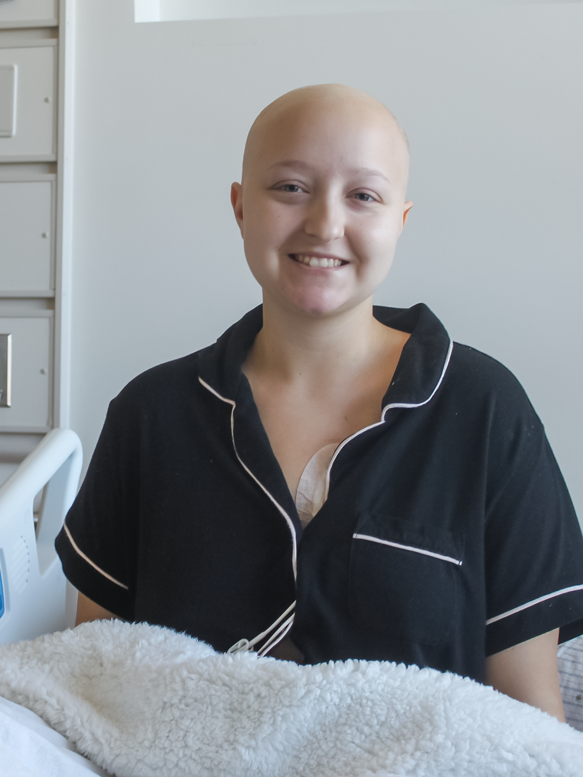 Woman with shaved head, smiling, wearing black pajamas, in a hospital bed.