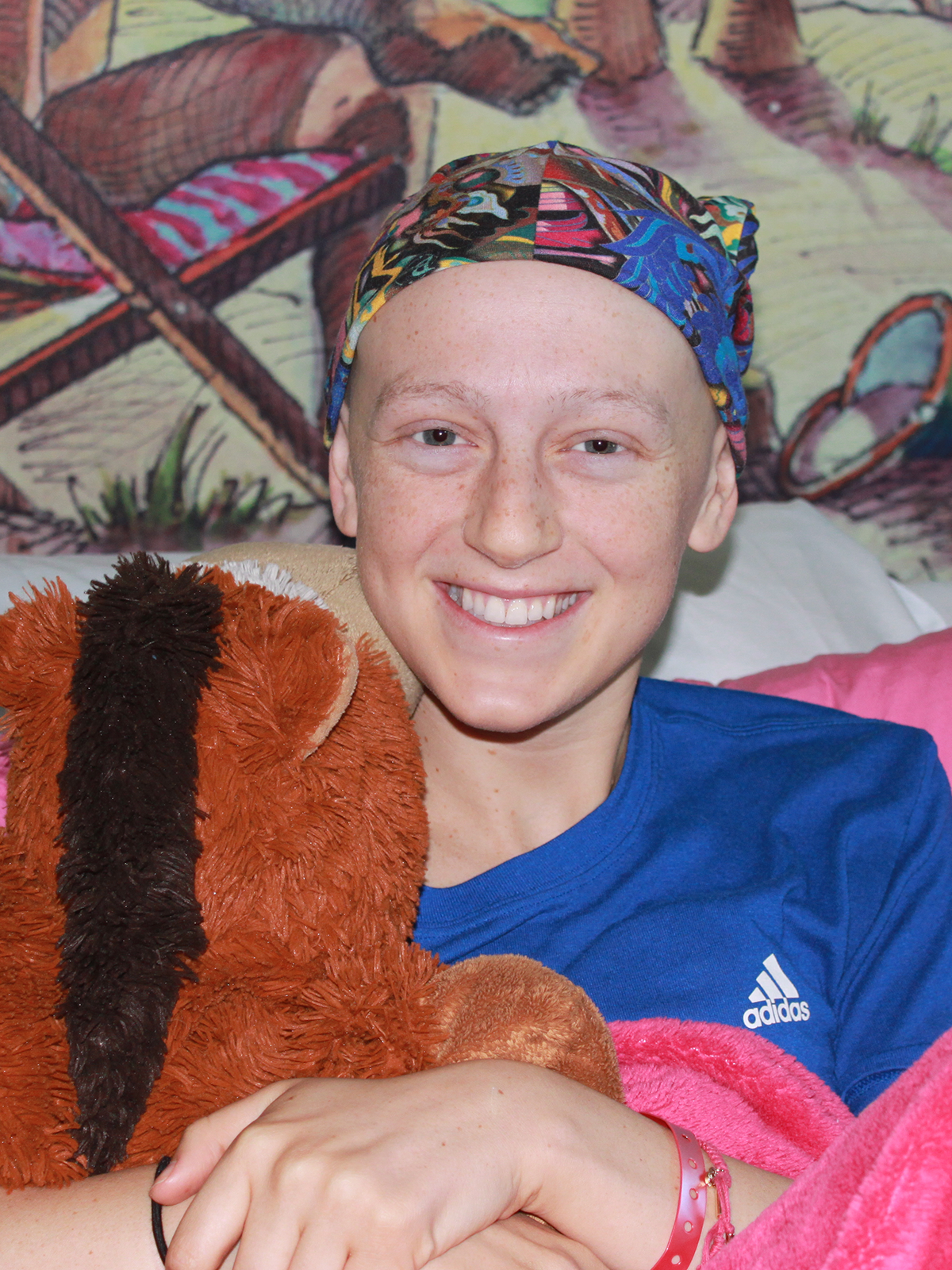 Smiling young woman with bald head wearing a patterned cap, holding a teddy bear.