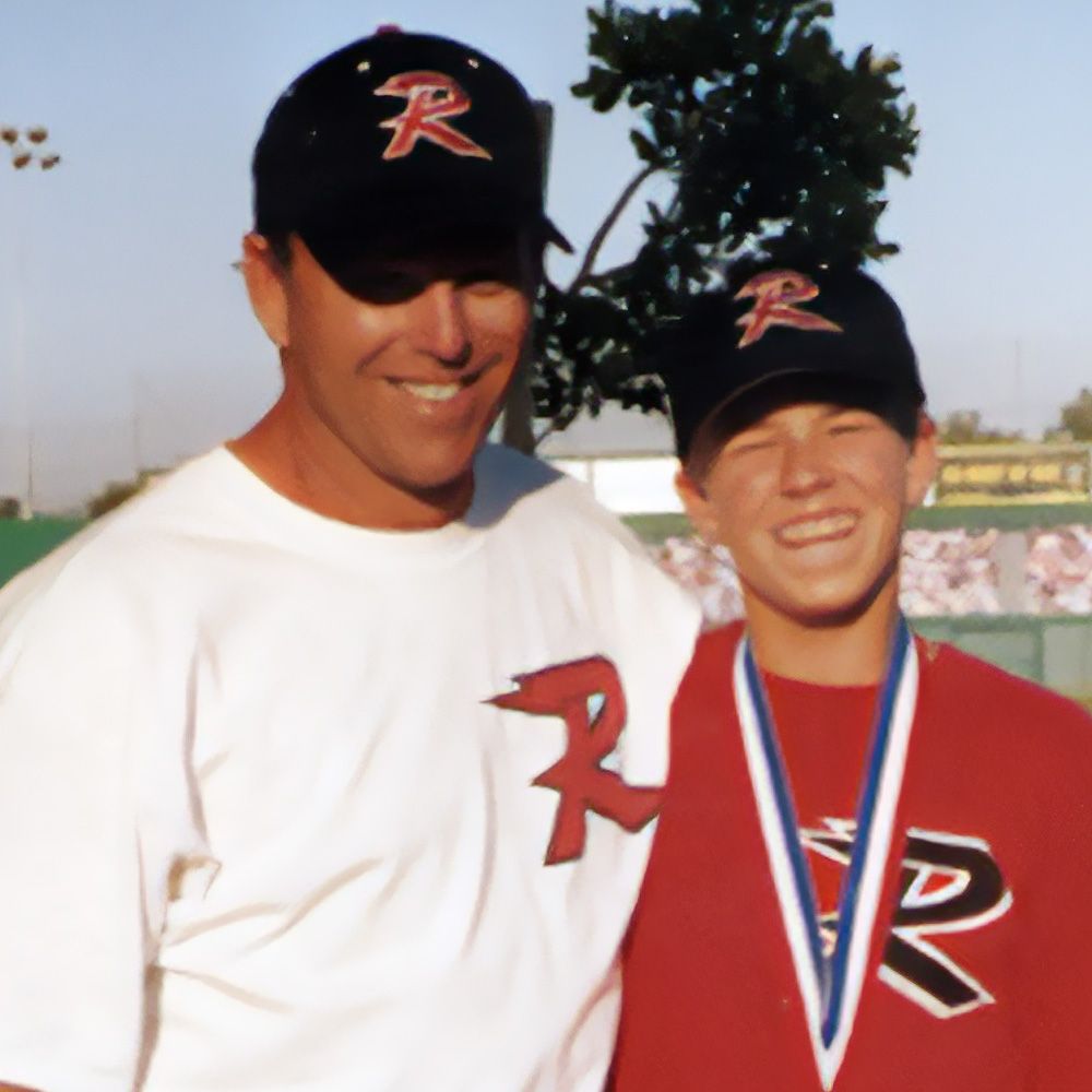 Man and boy in baseball hats and shirts, smiling, standing outdoors. The shirts have a large 