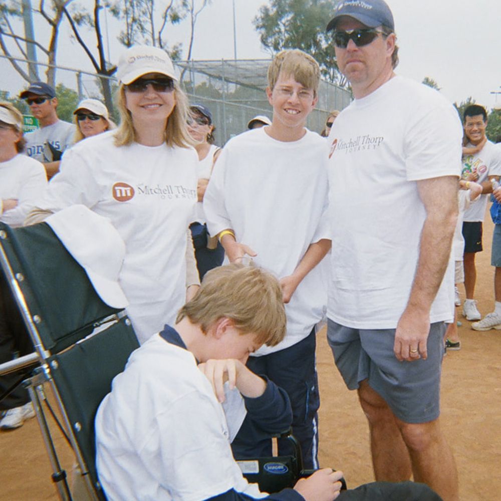 Family, including a boy in a wheelchair, pose outdoors at an event. They are wearing white shirts.