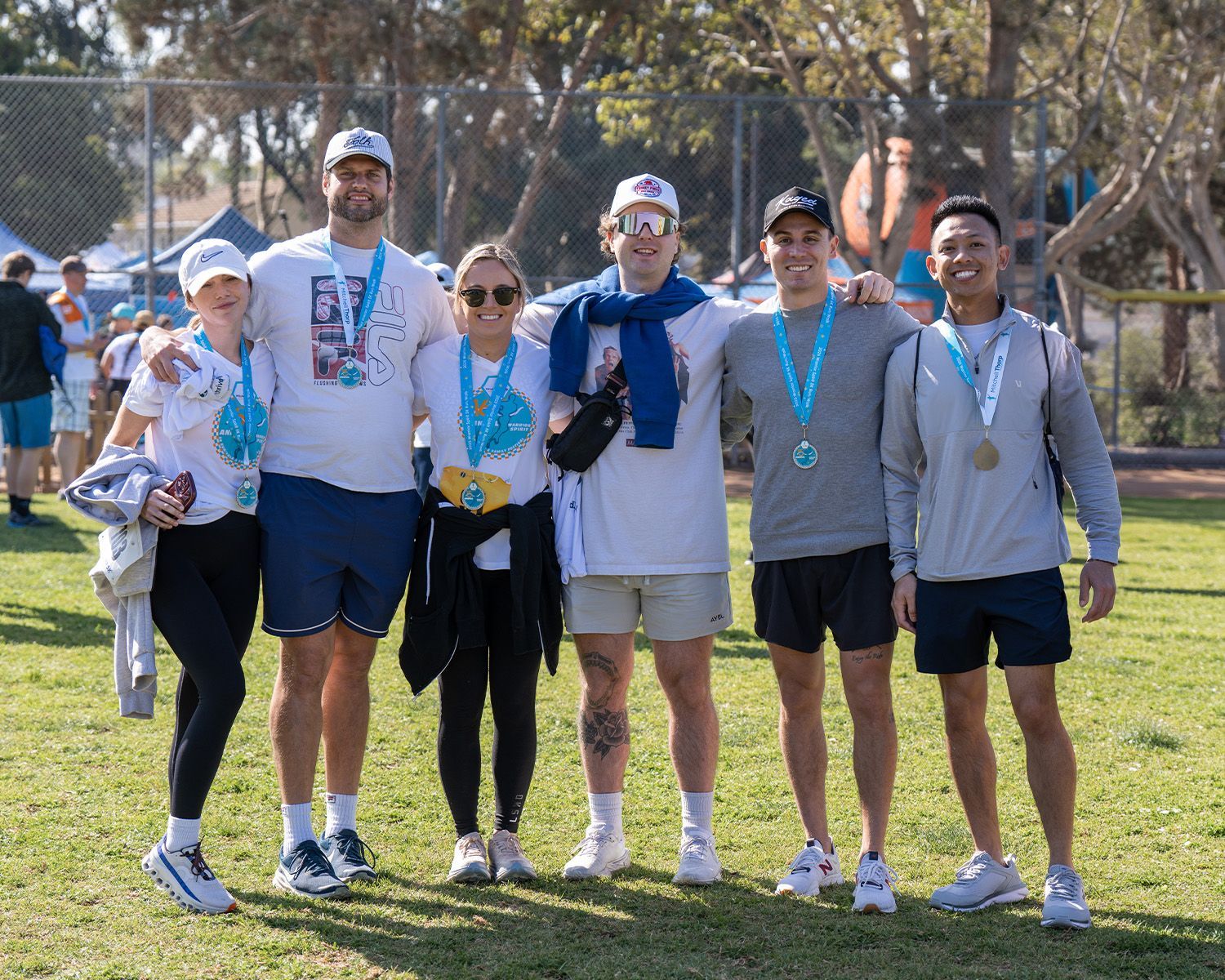 A group of six people stand together outdoors on grass wearing event medals, athletic clothing, and baseball caps.