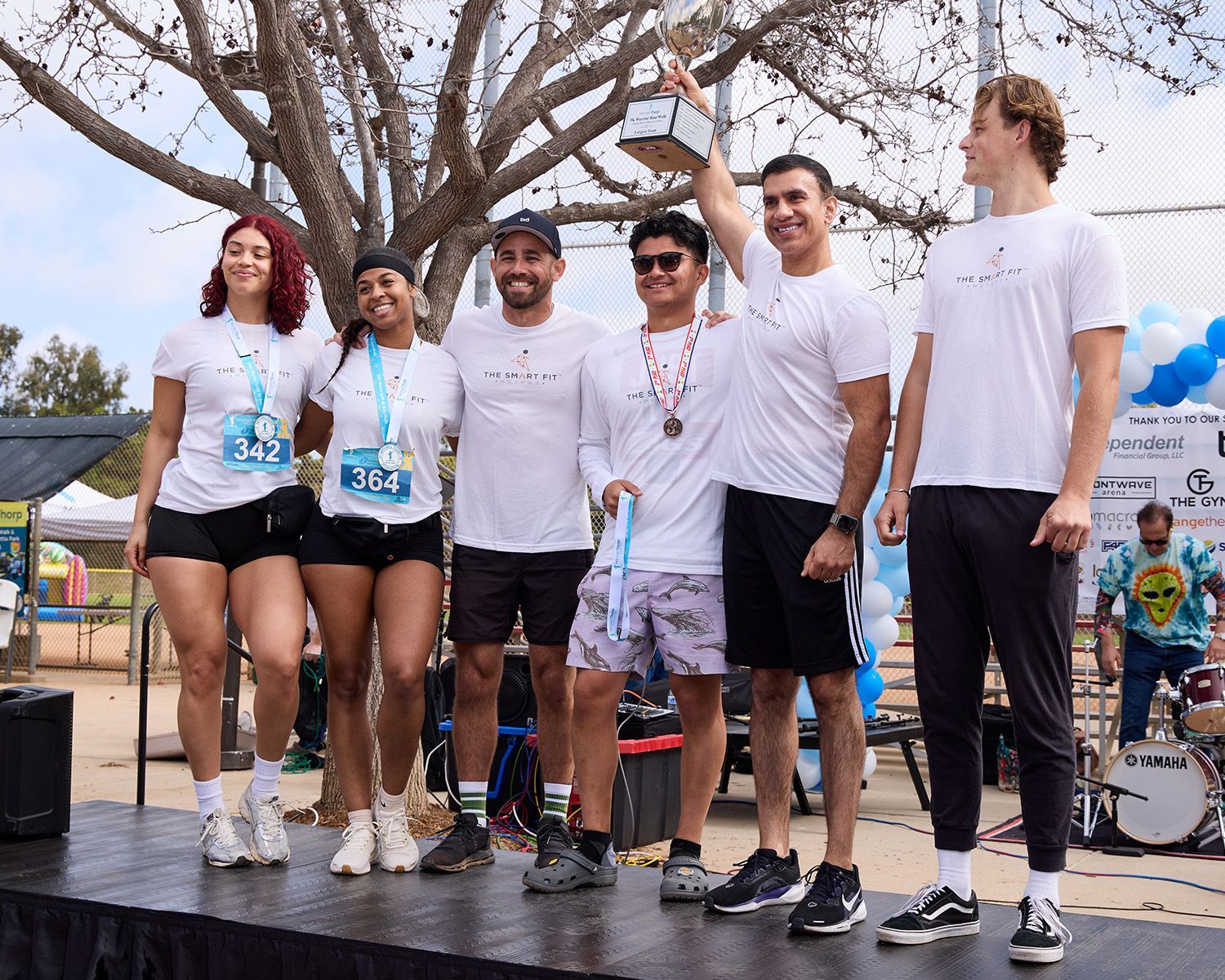 A group of six people stand together outdoors on grass wearing event medals, athletic clothing, and baseball caps.
