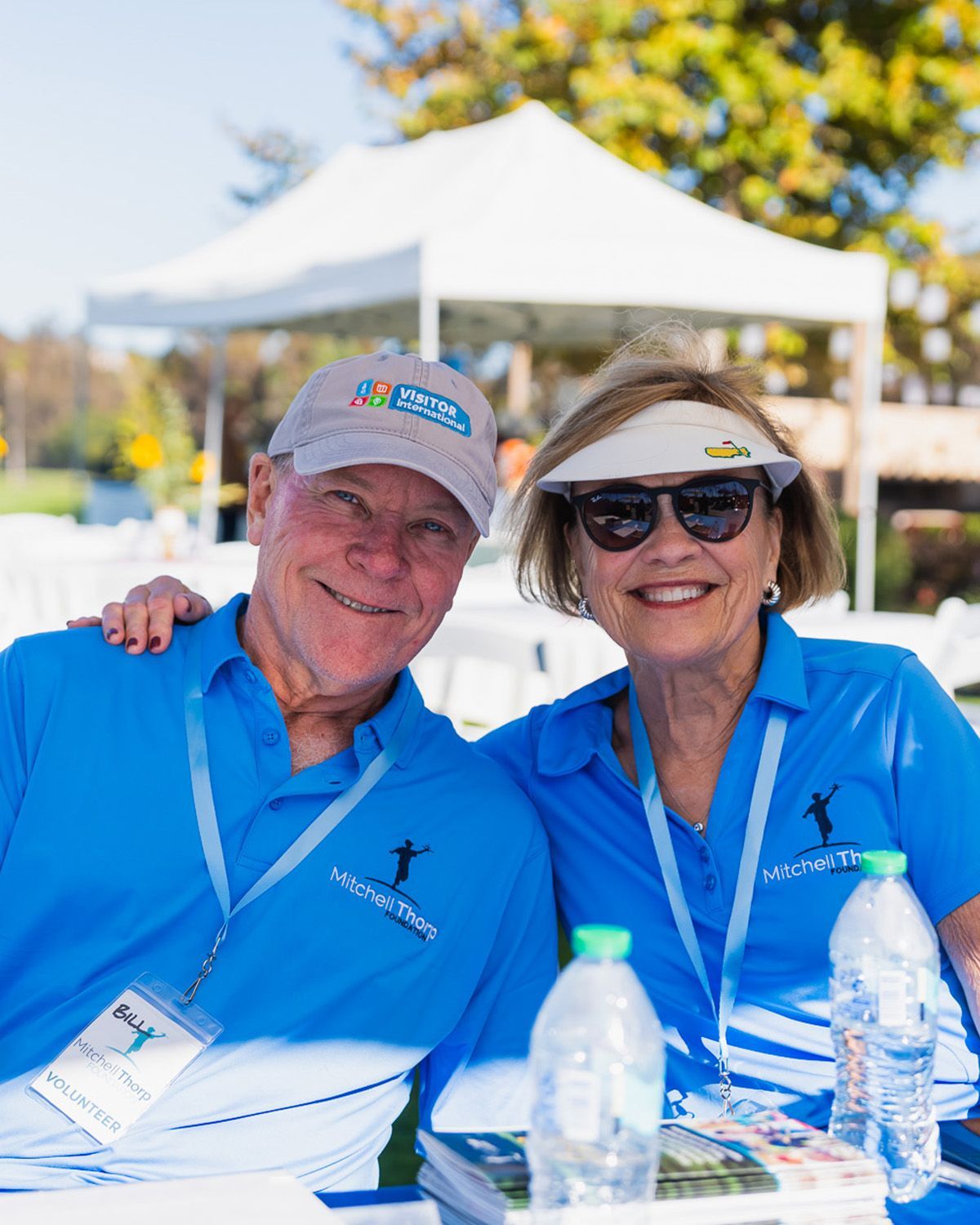 A man and woman wearing matching blue shirts and event lanyards smile together at an outdoor event under a white tent.