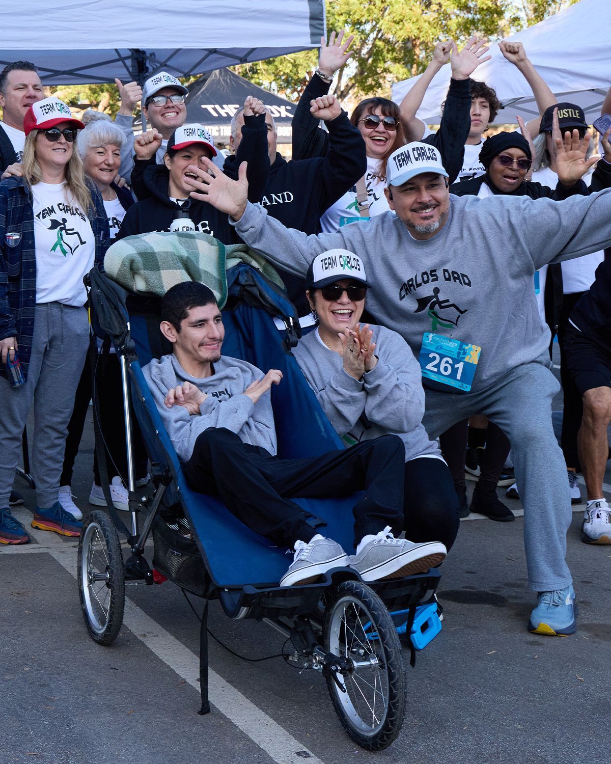 A group wearing matching hats and sweatshirts poses for a photo around a person in a racing wheelchair at an outdoor event.