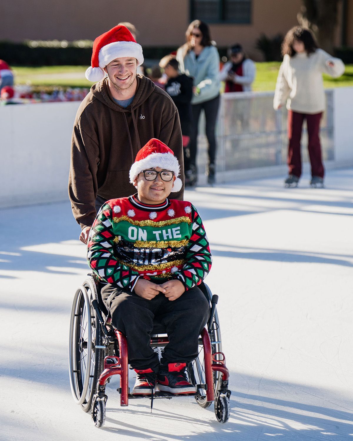 A person in a wheelchair wearing a festive sweater and Santa hat being pushed by another person on an ice rink.