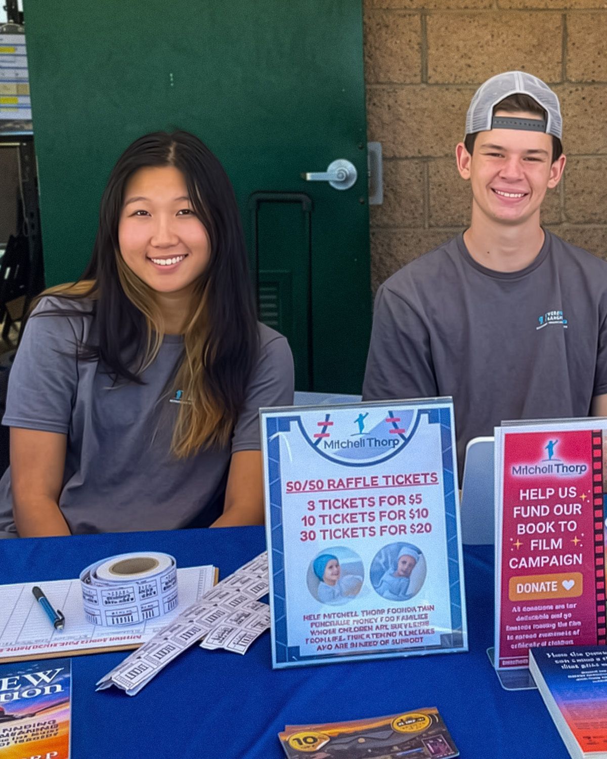 Two people sit at a table with a raffle ticket sign, promoting a campaign to fund a film project.