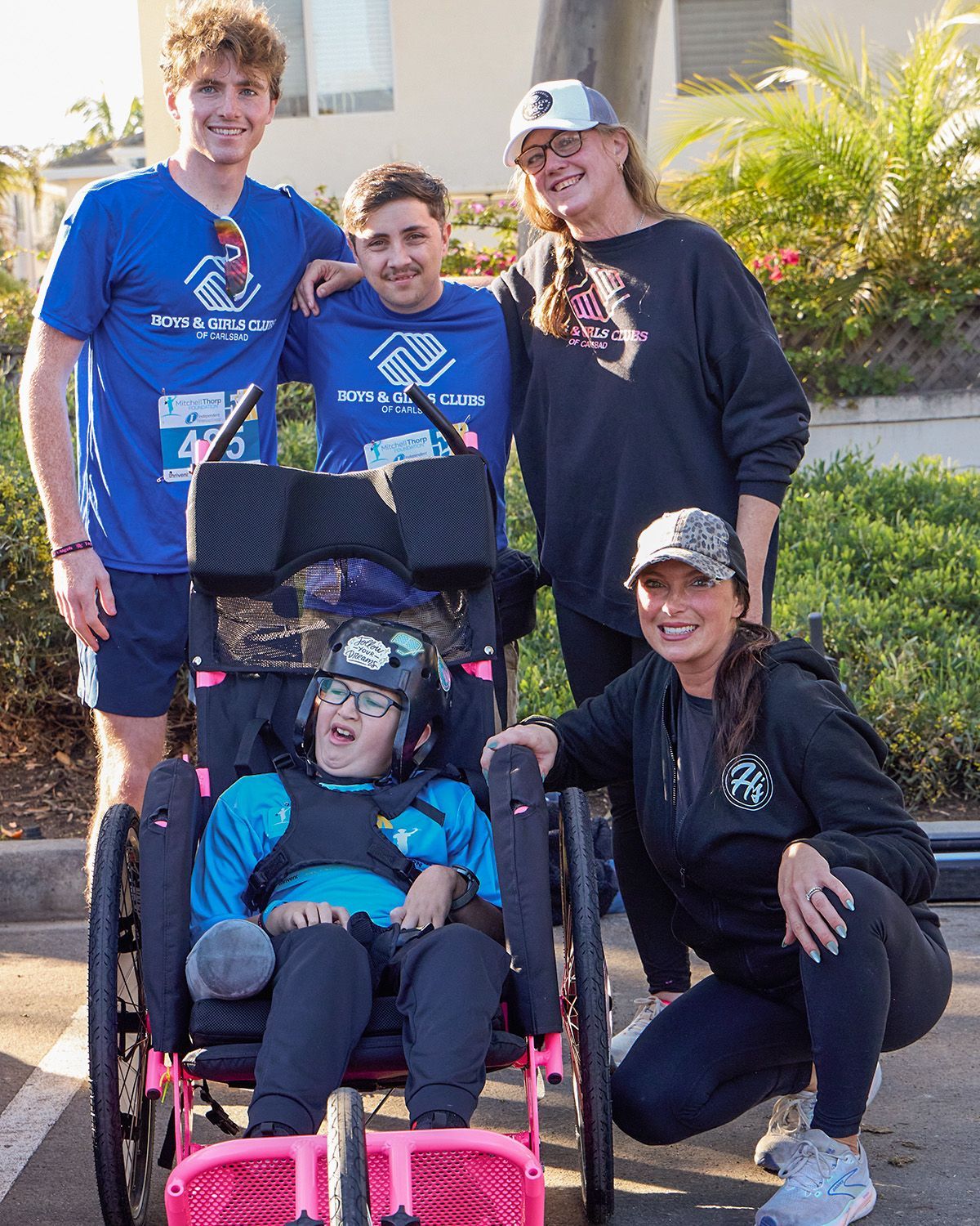 Four people pose outdoors, with one in a bright pink wheelchair. Everyone is smiling while wearing matching event shirts.