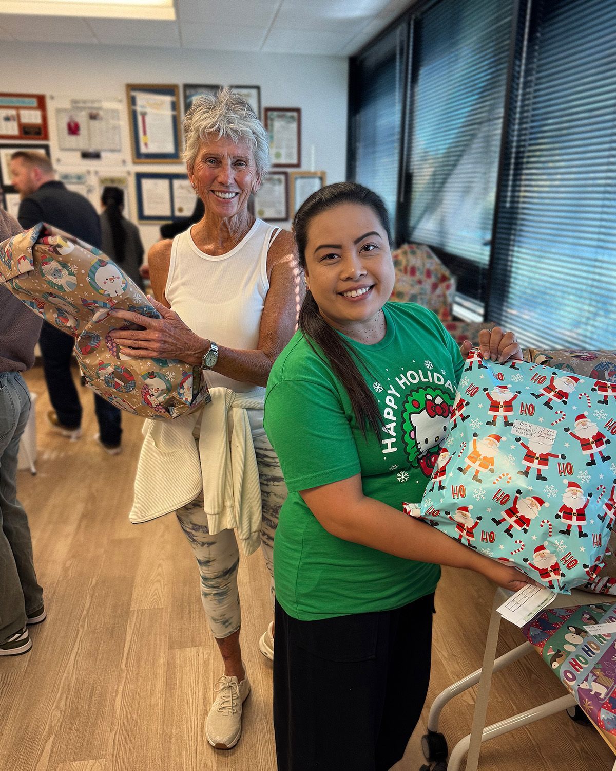 Two people standing in an office holding wrapped Christmas gifts, one wearing a green t-shirt and the other a white tank.