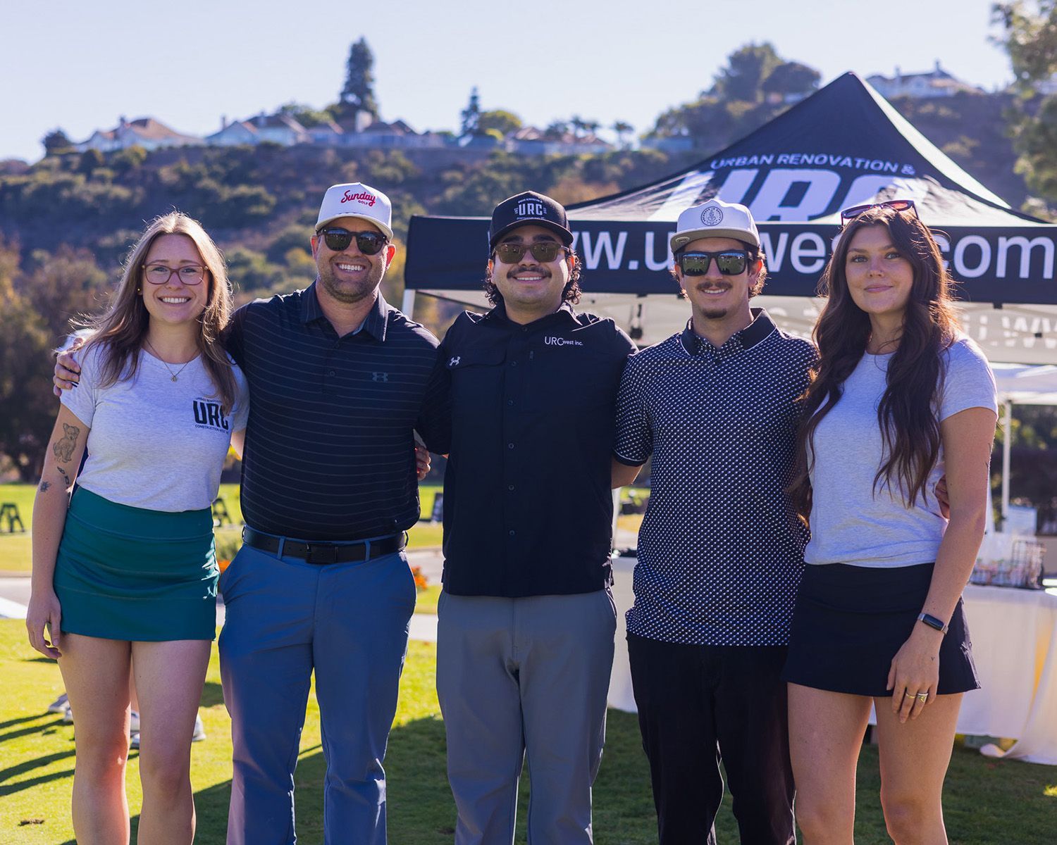 Five people stand outdoors in front of a branded tent at a golf course, smiling for a group photo on a sunny day.