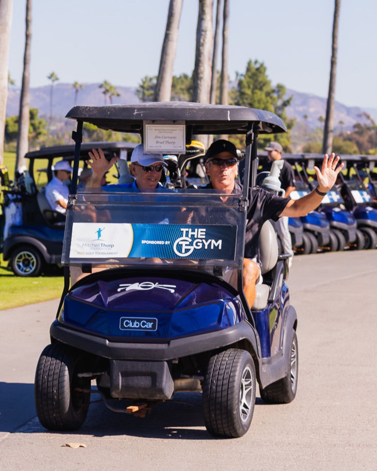 Two men in a blue golf cart wave while driving down a sunny path lined with palm trees and other golf carts.