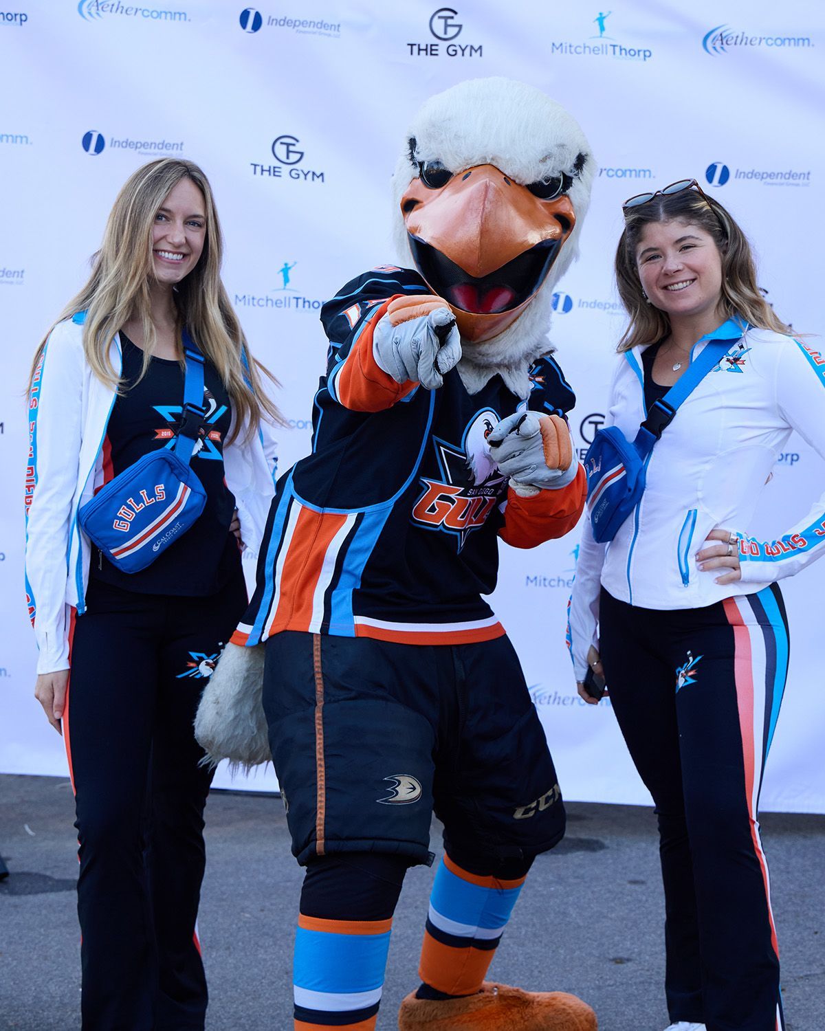 Two people stand on either side of a mascot in a sports uniform, pointing toward the viewer against a branded backdrop.