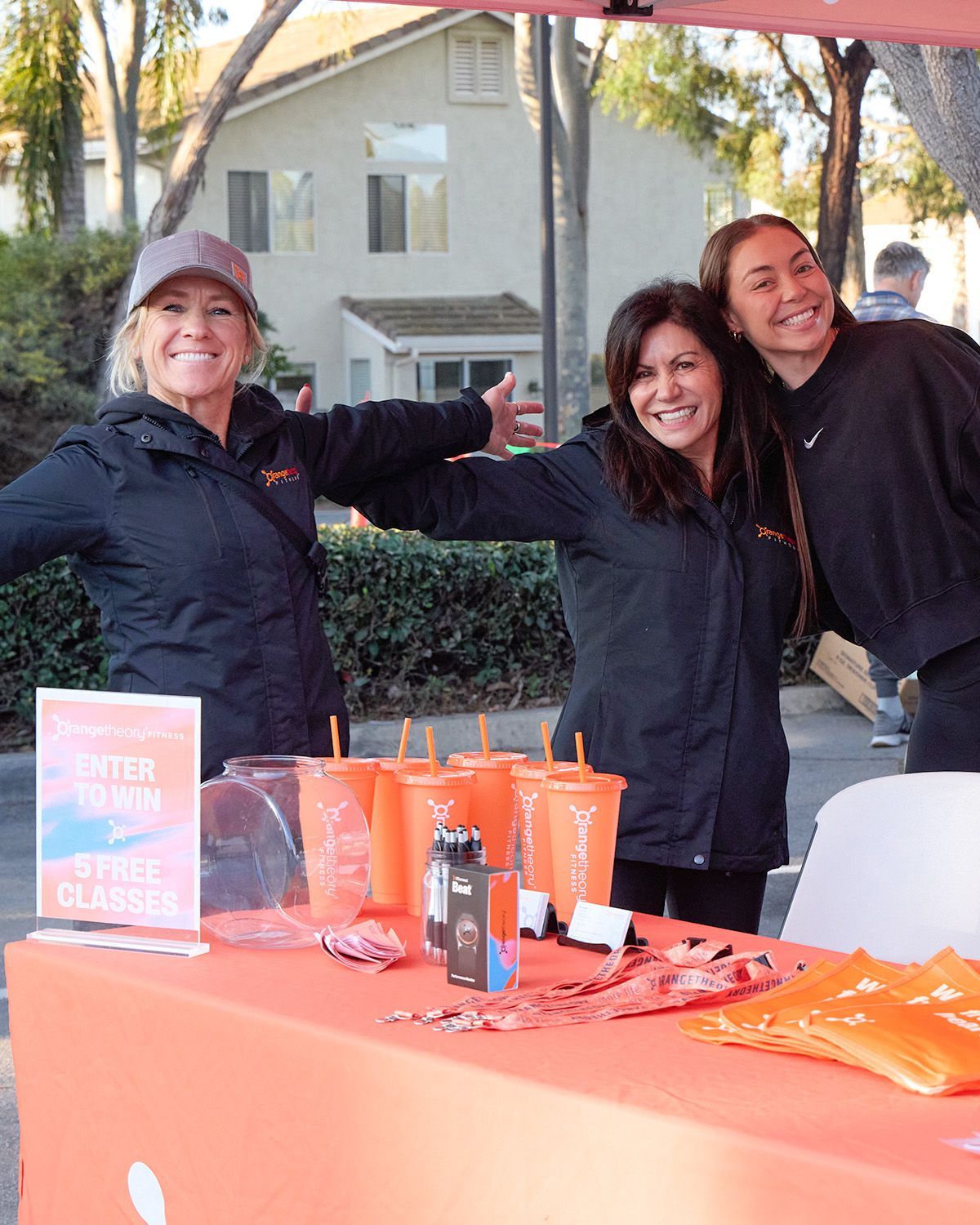 Three people smiling behind an orange event table with orange cups and flyers in front of a building.