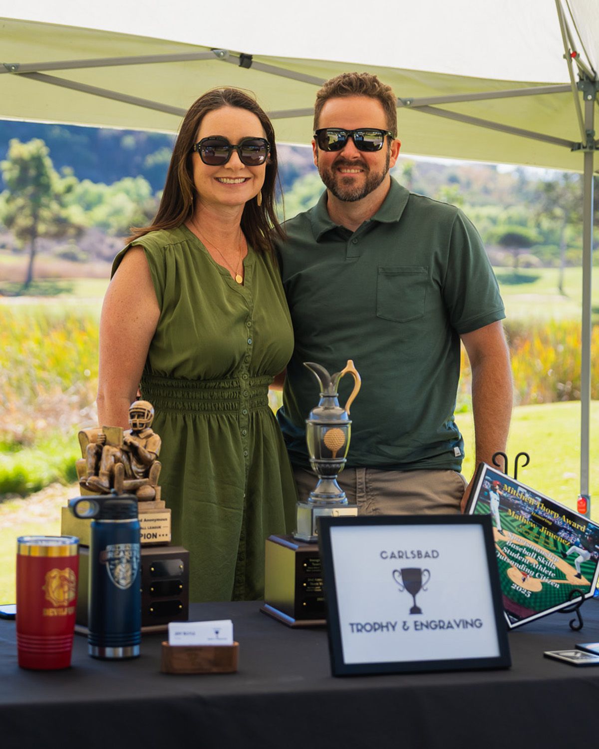 A person in a green dress and a person in a green shirt stand behind a table with trophies, outdoors at a golf course.