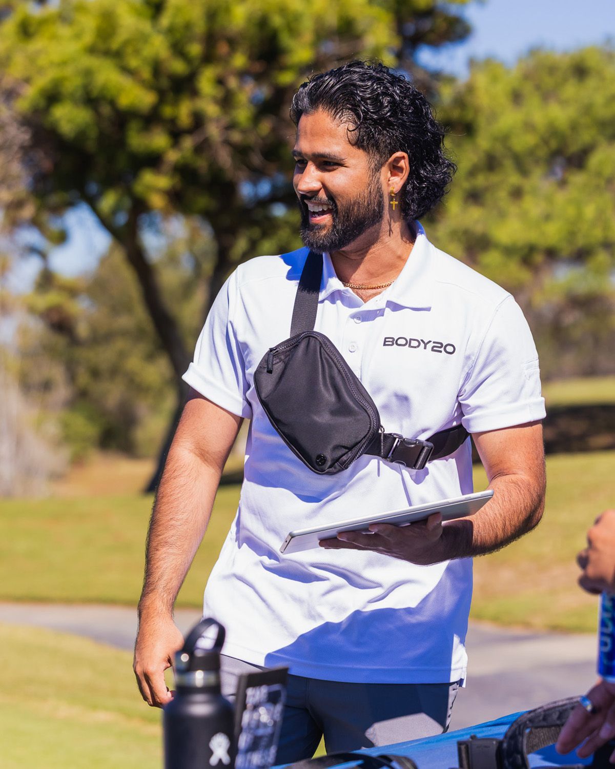 A smiling person in a white collared shirt and black crossbody bag holds a tablet outdoors on a sunny day.