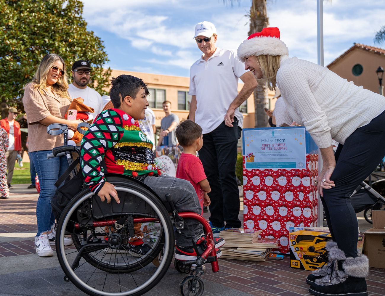 Boy in wheelchair receives gift from woman in Santa hat at outdoor event.