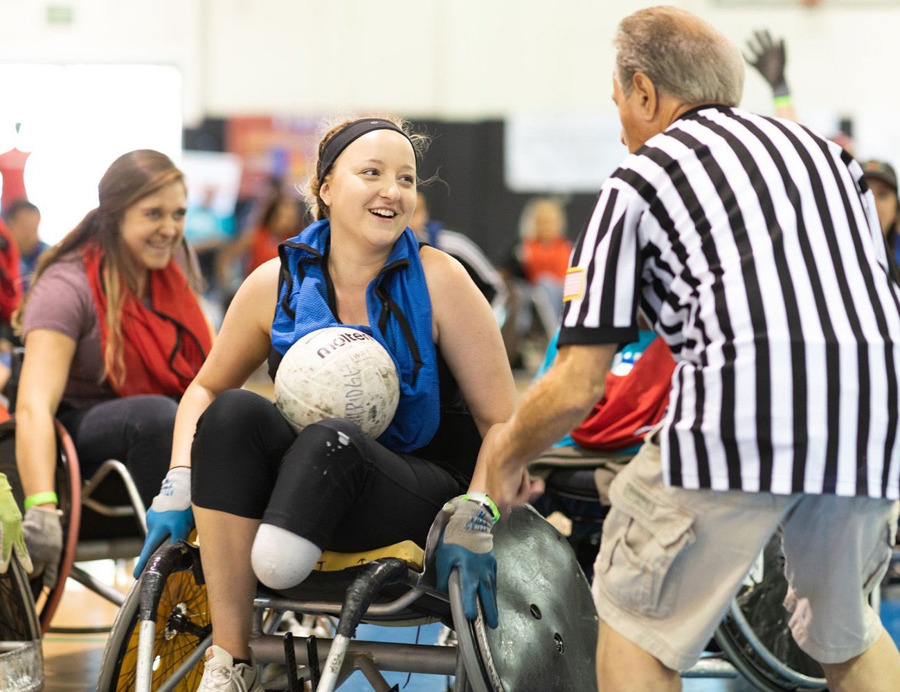 Woman in wheelchair smiles, holding ball, interacting with referee at a sports event.
