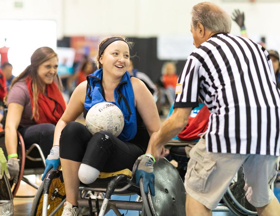 Woman in wheelchair smiles, holding ball, interacting with referee at a sports event.