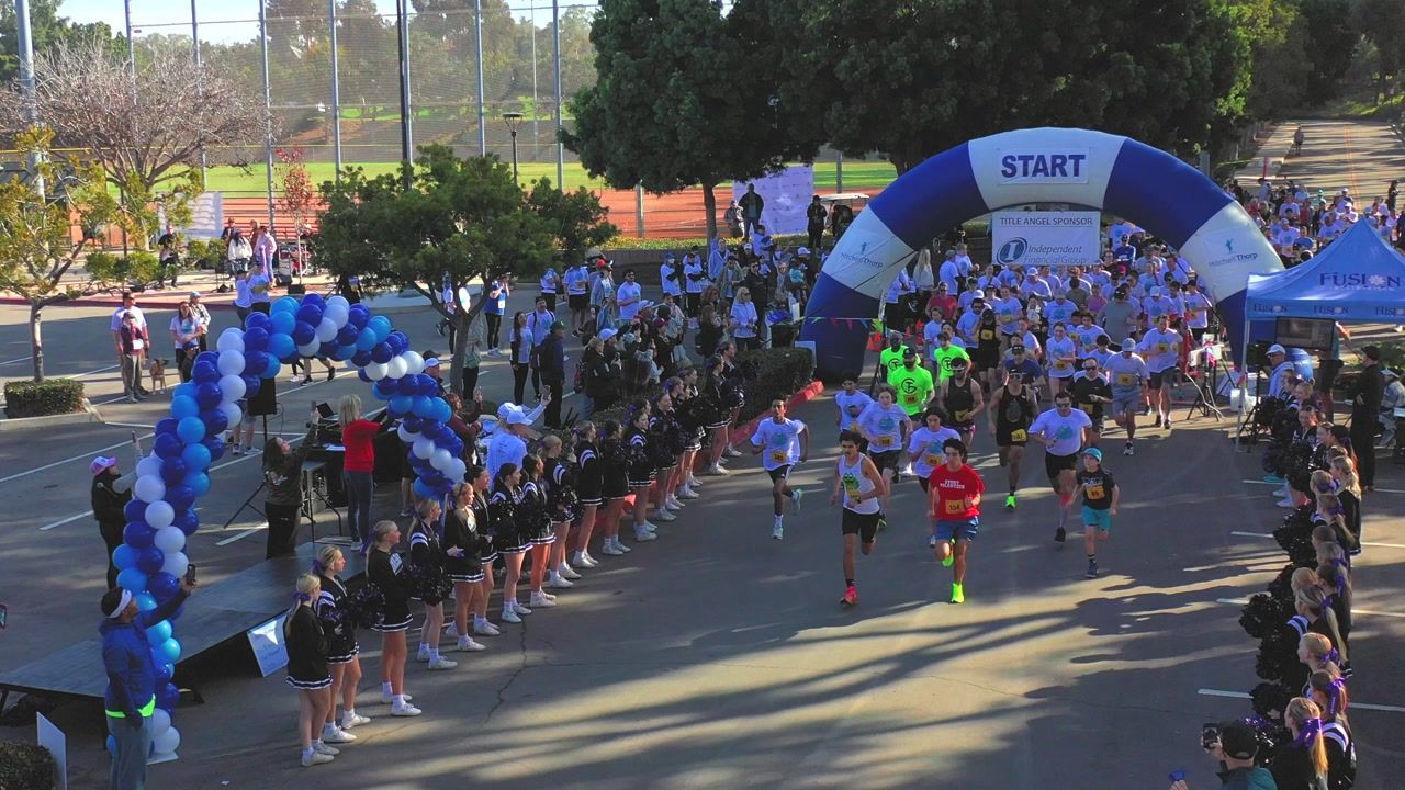 Runners starting a race under a blue and white inflatable arch, lined with supporters; sunny outdoor setting.