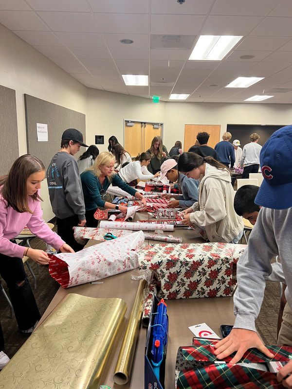 People wrapping gifts at a long table. Wrapping paper rolls are visible. Room is brightly lit.