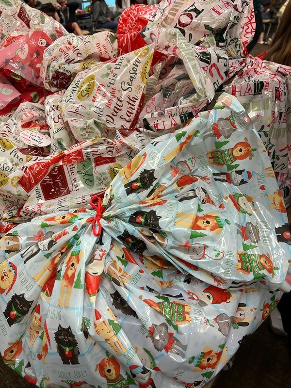 Pile of wrapped gifts in various festive wrapping papers, tied with red ribbons.