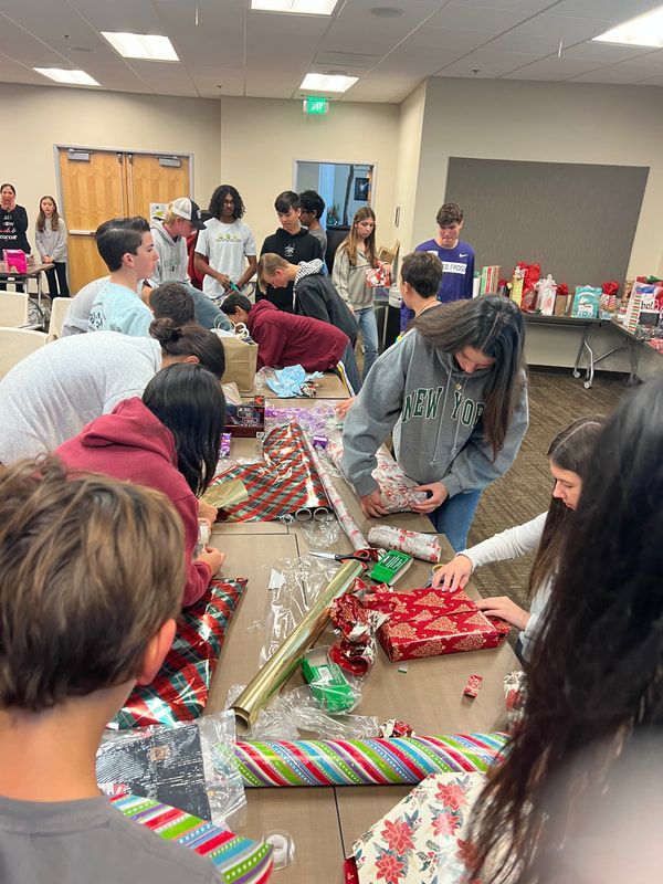 A group of people wrapping gifts at a table.  Wrapping paper and presents are on the table.