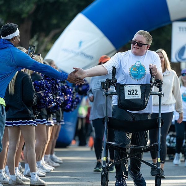 Person using walker high-fives a man at a race finish line with cheerleaders in the background, under an arch.