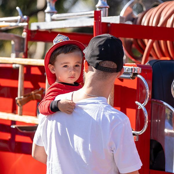 Boy in firefighter hat on a person's shoulders, near a fire truck. The boy looks at the camera.
