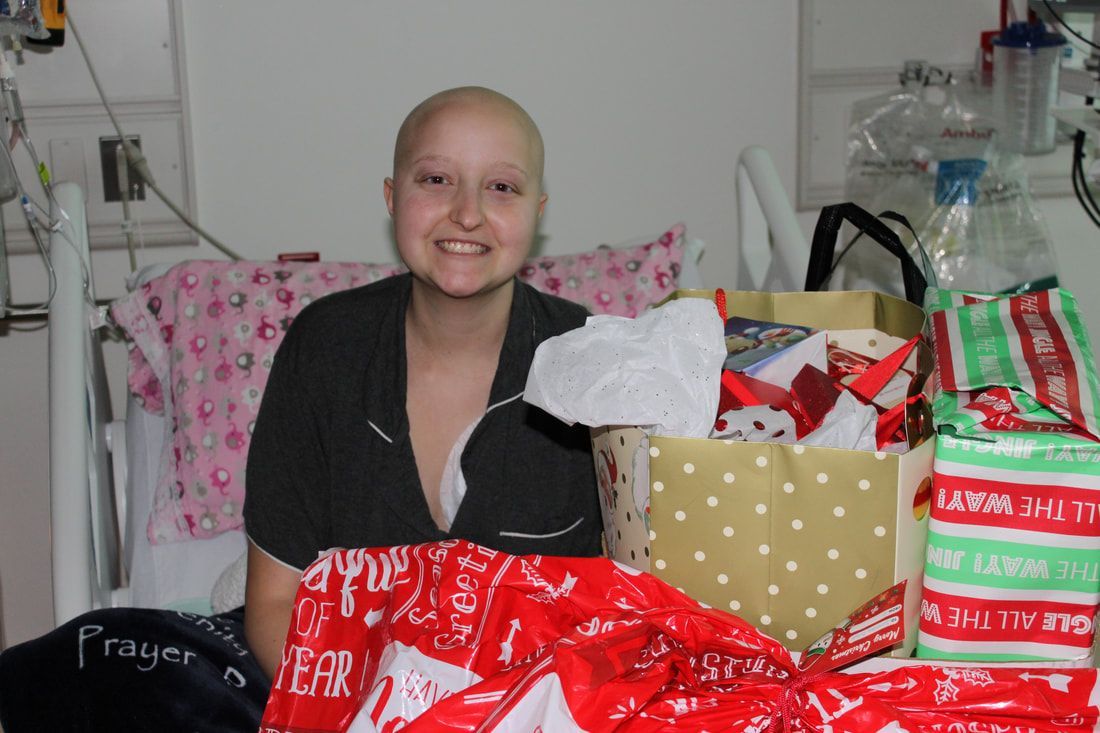 Smiling person with bald head, surrounded by wrapped gifts in a hospital bed.